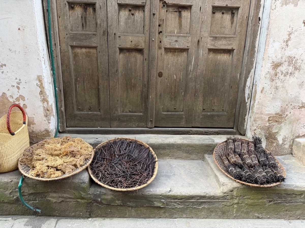 Woven trays filled with dried vanilla pods and island aromatics outside a weathered Zanzibar doorway