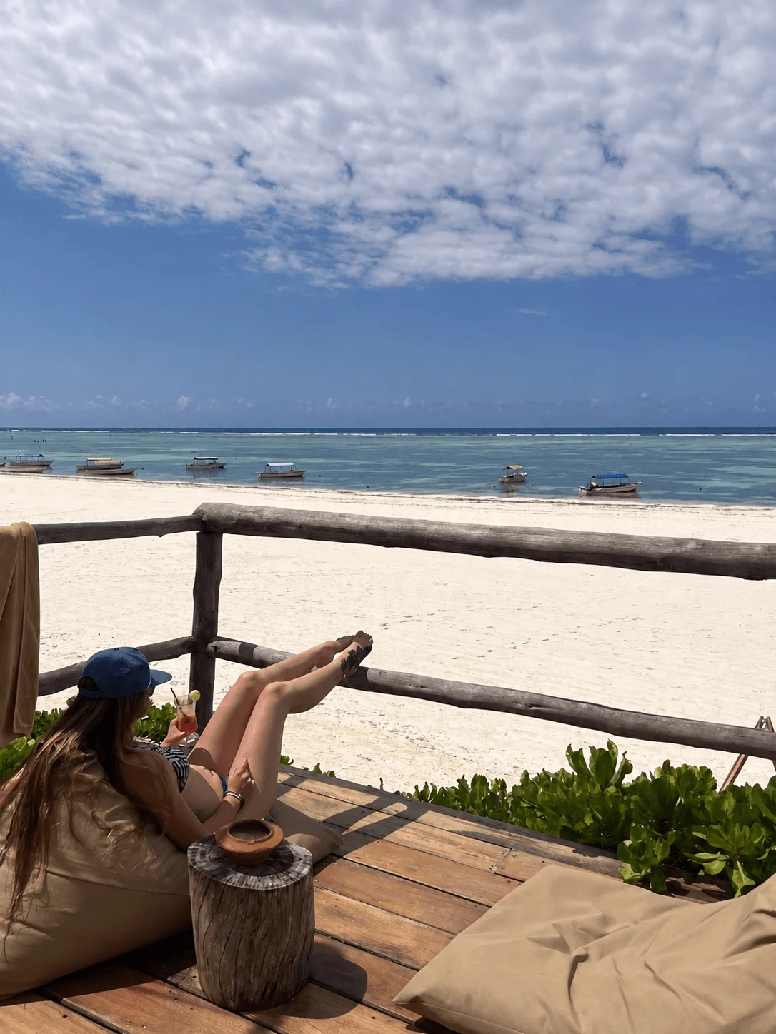 A traveler relaxing with a drink overlooking a white beach, turquoise water, and anchored boats